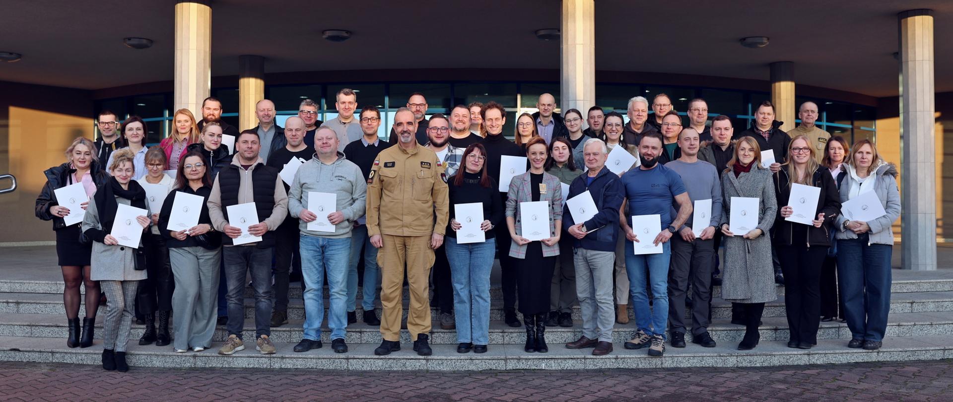 Pamiątkowe zdjęcie grupowe uczestników szkolenia inspektorów ochrony przeciwpożarowej (41 osób) trzymających w dłoniach otrzymane białe teczki w towarzystwie Zastępcy Komendanta Centralnej Szkoły PSP, kobiety oraz funkcjonariusza CS PSP