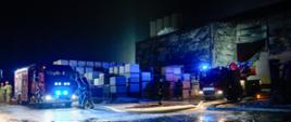 Na zdjęciu widać nocną akcję strażacką na terenie zakładu przemysłowego. Na mokrym, oblodzonym placu stoją dwa wozy strażackie z włączonymi niebieskimi światłami ostrzegawczymi, których refleksy odbijają się od nawierzchni. Strażacy w ubraniach ochronnych poruszają się między pojazdami i rozwiniętymi wężami gaśniczymi. W tle znajduje się duża hala magazynowa z uszkodzoną elewacją, obok której ułożone są wysokie stosy bloczków lub palet.
