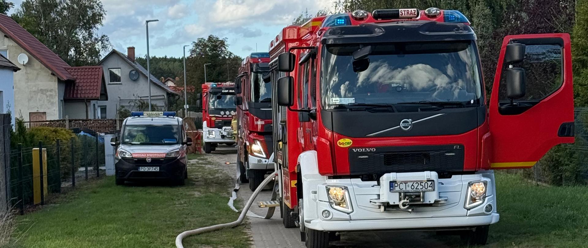 Wóz strażacki z rozłożoną linią gaśniczą podłączoną do hydrantu na drodze osiedlowej, w tle kolejne pojazdy PSP i budynki mieszkalne.