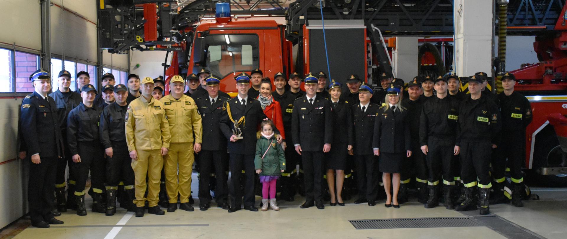 Zdjęcie strażaka odchodzącego na zaopatrzenie emerytalne. Trzyma on w rekach pamiątkową statuetkę. Po jego lewej stronie jest jego córka i żona. Pozują oni do zdjęcia grupowego wraz z pracownikami KP PSP w Skarżysku-Kamiennej na tle drabiny pożarniczej.