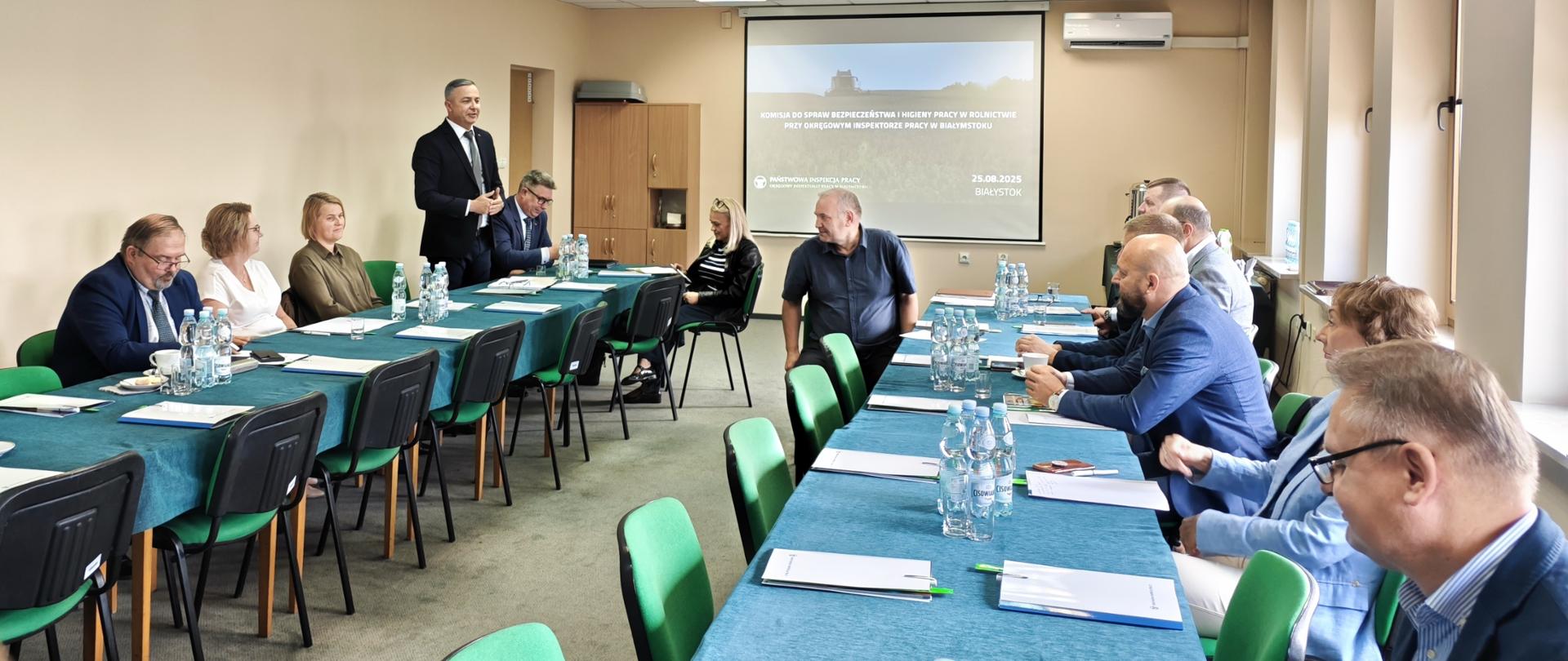 Uczestnicy posiedzenia siedzą przy stołach konferencyjnych. Jeden z mężczyzn stoi i wygłasza przemówienie. W tle znajduję się ekran z prezentacją.