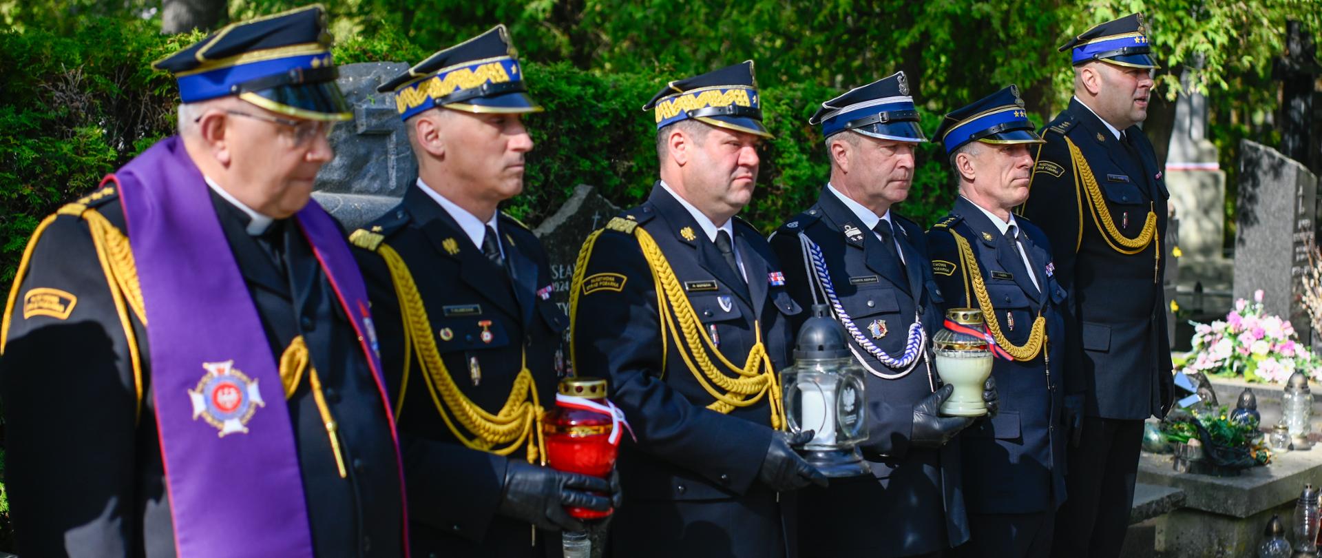 Grupa strażaków w galowych mundurach Państwowej Straży Pożarnej oraz ksiądz kapelan stoją w rzędzie na cmentarzu podczas uroczystości, trzymając znicze.