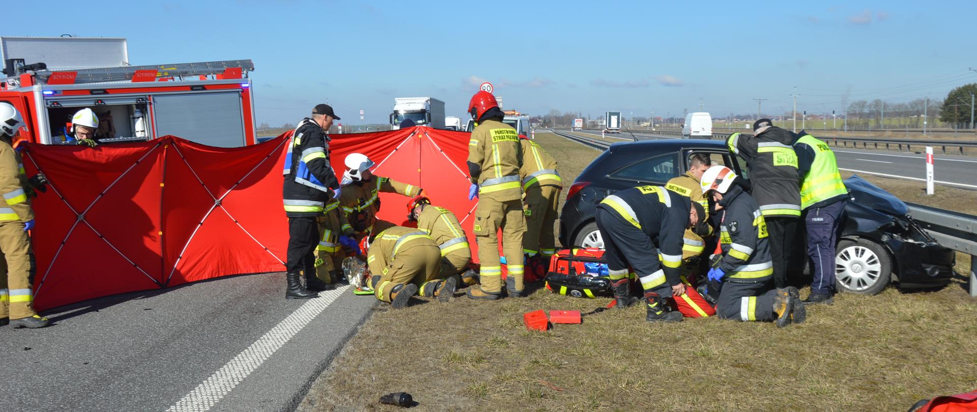 Zderzenie pojazdów na S5. Odcinek ekspresowej "piątki" pomiędzy węzłami Bojanowo i Rawicz. Na pasie zieleni strażacy udzielają kwalifikowanej uczestniczkom wypadku. Jedna z nich leży na pasie zieleni, druga przebywa w samochodzie. Miejsce prowadzenia działań zabezpieczone jest przy pomocy czerwonego parawanu. Za nim widoczny jest fragment wozu strażackiego.