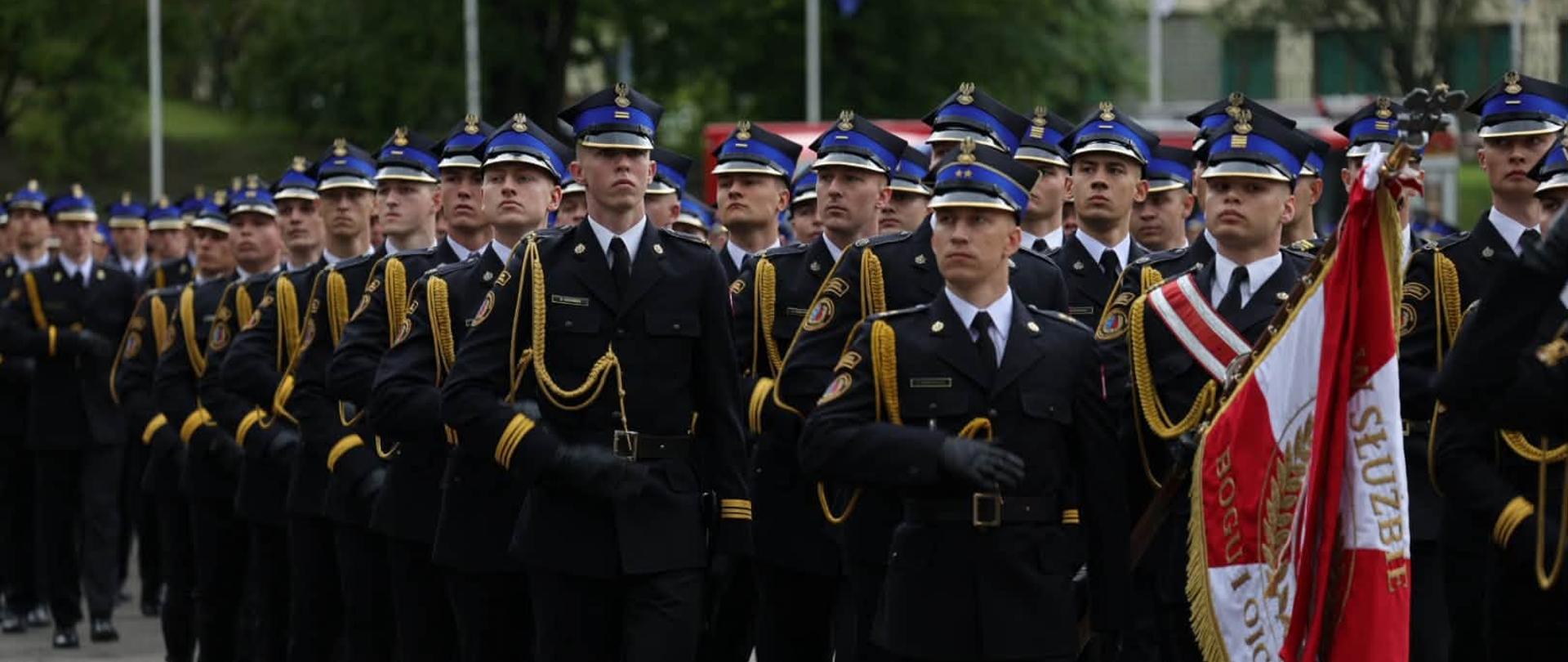 10akademia.jpg – Strażacy maszerujący w zwartym szyku podczas uroczystości. Na czele pododdziału niesiony jest biało-czerwony sztandar.