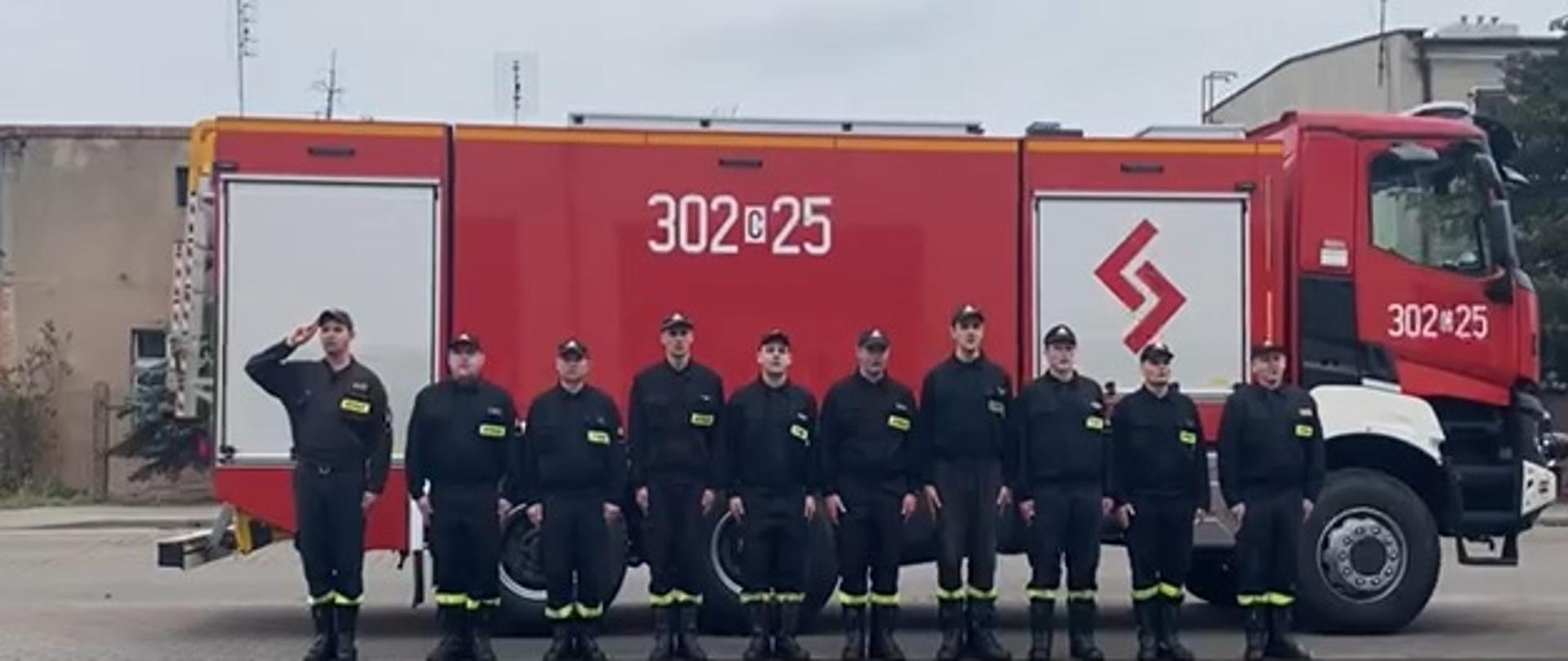 Zbiórka na placu zewnętrznym JRG2 w Bydgoszczy, strażacy w szeregu w czarnych ubraniach koszarowych, za nimi stojący bokiem czerwony samochód pożarniczy. 