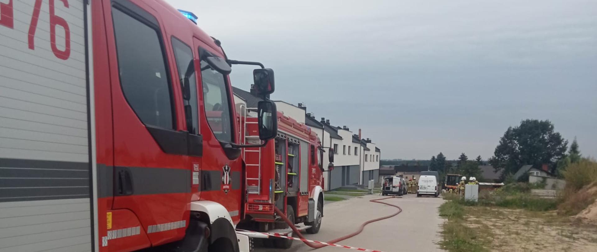 Na zdjęciu pojazdy straży pożarnej. Zdjęcie wykonane w ciągu dnia. Teren budowanego osiedla. Zachmurzenie duże. W oddali radiowóz Policji. Miejsce wygrodzone.