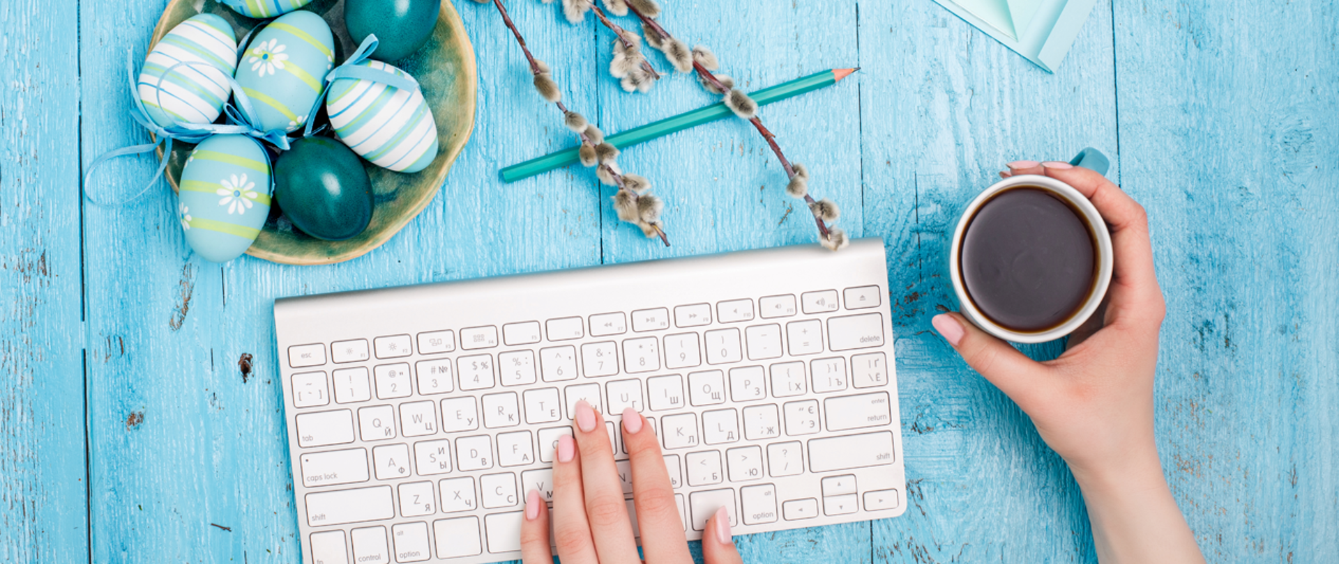 Na drewnianym niebieskim stole dłoń na klawiaturze komputera po prawej stronie ręka trzymająca kubek z kawą u góry ozdoby wielkanocne w niebieskich barwach