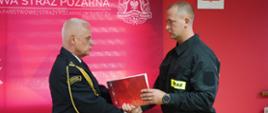Świetlica komendy. Na tle ścianki promocyjnej zastępca komendanta powiatowego, występujący w umundurowaniu wyjściowym ze sznurem galowym, składa gratulacje i wręcza teczkę z aktem ślubowania nowo przyjętemu do słuzby strażakowi. 