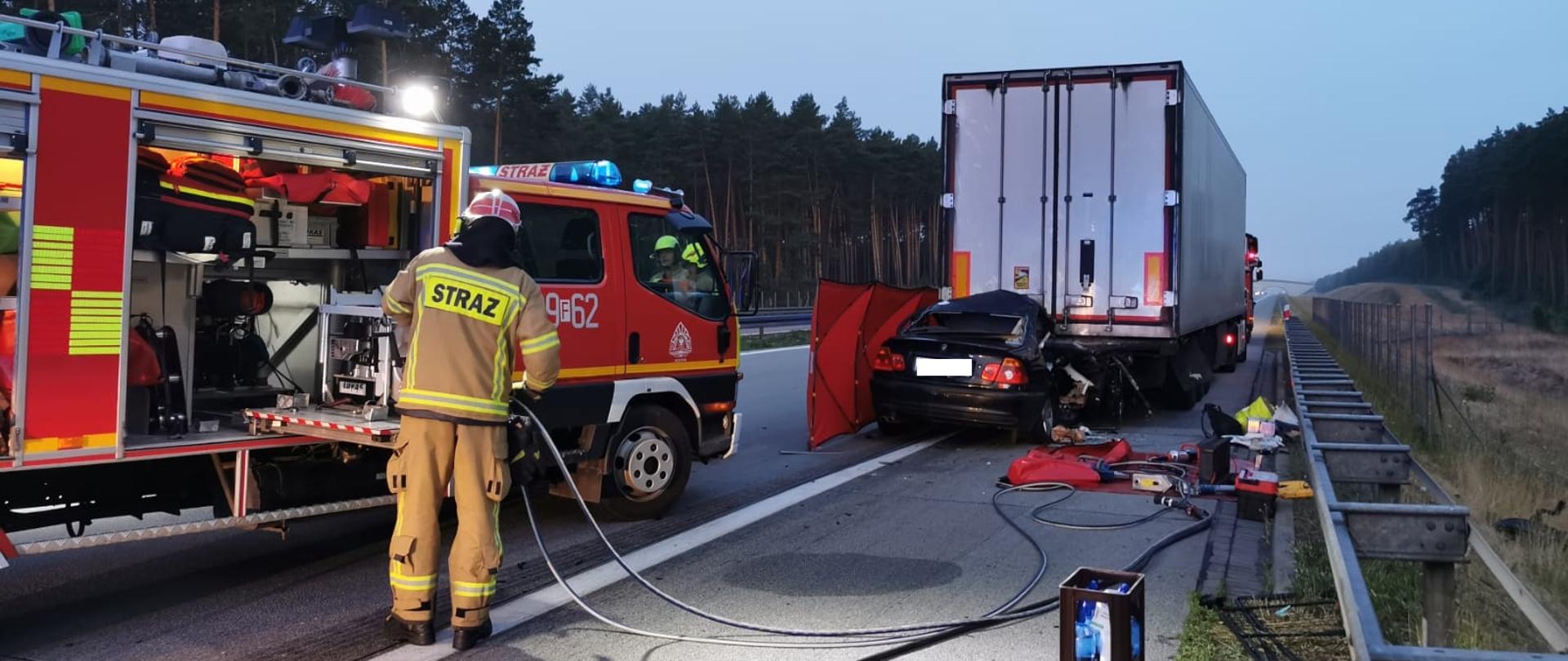 Stojący na pasie awaryjnym autostrady samochód ciężarowy z naczepą. W tył naczepy wbity samochód osobowy. Na jezdni na macie rozłożone narzędzia ratownicze straży pożarnej. Po lewej stronie samochód strażacki i obok umundurowany strażak.