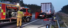 Stojący na pasie awaryjnym autostrady samochód ciężarowy z naczepą. W tył naczepy wbity samochód osobowy. Na jezdni na macie rozłożone narzędzia ratownicze straży pożarnej. Po lewej stronie samochód strażacki i obok umundurowany strażak.