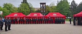 Zdjęcie przedstawia poczty sztandarowe straży pożarnych. Na drugim planie widać czerwone namioty z logo Państwowej Straży Pożarnej. 