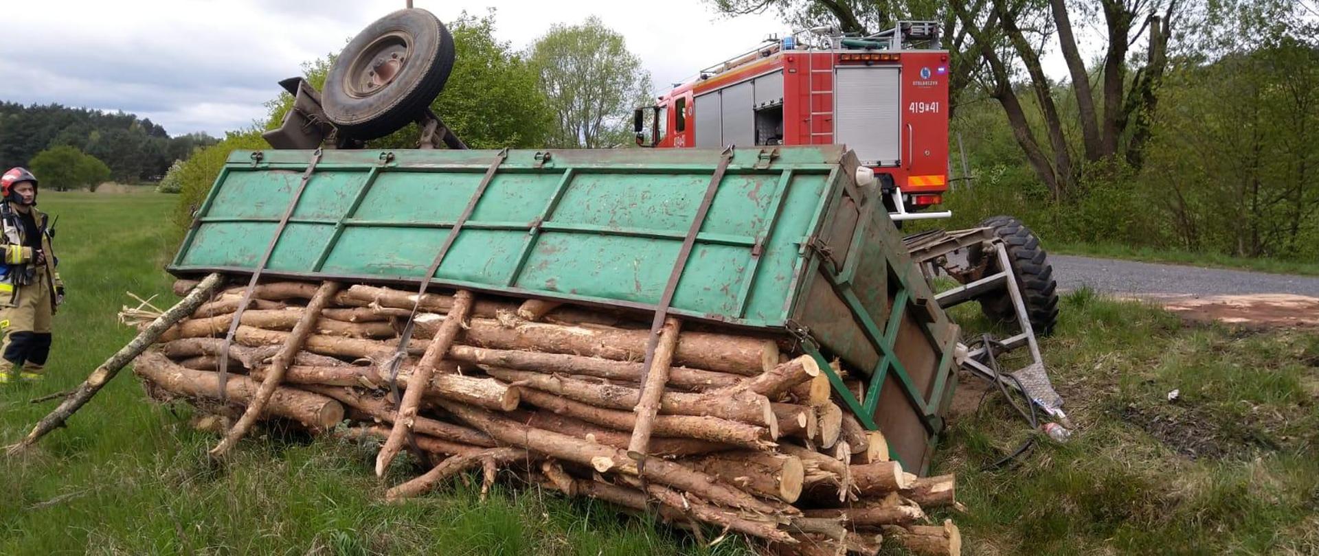 wywrócona przyczepa z drewnem. W tle samochód strażacki