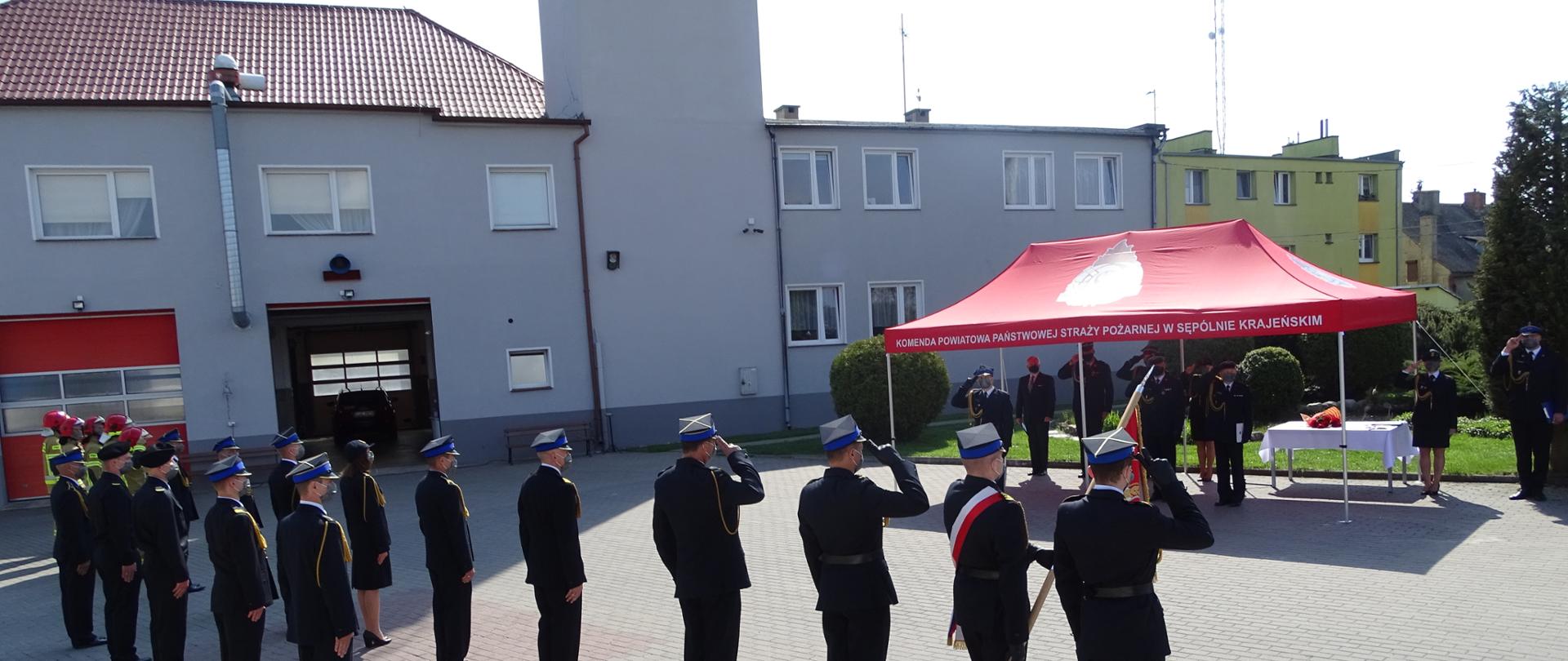 Zdjęcie wykonano podczas uroczystości Obchodów Dnia Strażaka w Komendzie Powiatowej Państwowej Straży Pożarnej w Sępólnie Krajeńskim.
Na zdjęciu widzimy pododdziały Komendy Powiatowej Państwowej Straży Pożarnej w Sępólnie Krajeńskim podczas podnoszenia Flagi Państwowej. Na pierwszym planie poczet sztandarowy i pododdziały Komendy Powiatowej Państwowej Straży Pożarnej w Sępólnie Krajeńskim, w tle pod czerwonym namiotem Komendy Powiatowej Państwowej Straży Pożarnej w Sępólnie Krajeńskim Panowie Komendanci oraz zgromadzeni goście. W tle widoczny budynek Komendy Powiatowej Państwowej Straży Pożarnej w Sępólnie Krajeńskim. Wszyscy uczestnicy ceremonii mają na sobie maseczki jednorazowe.
