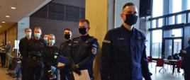 Na zdjęciu widzimy funkcjonariuszy, mężczyźni oraz kobiety stojący w kolejce. dwie pierwsze osoby to policjanci ubrani w granatowe mundury, na prawych ramionach napis policja, czarne maseczki na twarzy, czarne rękawiczki, w rękach trzymają białe kartki. Kolejne osoby w kolejce to strażacy ubrani w czarne mundury, niebieskie maski na twarzy, niebieskie rękawiczki. Całość dzieje się w pomieszczeniu, dalej z prawej strony kadru widzimy okna od podłogi aż po sufit, przeszklone drzwi wejściowe oraz czerwone krzesełka. Lewa dalsza strona kadru to osoby siedzące i stojące słabo widoczne również stojące w kolejce. Podłoga pomieszczenia koloru brązowego. 
