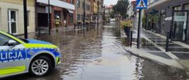 Zalana ulica miejska. Po lewej stronie stoi radiowóz policji. Na dalszym planie widać strażaków poruszających się w wodzie, która sięga do krawężników.