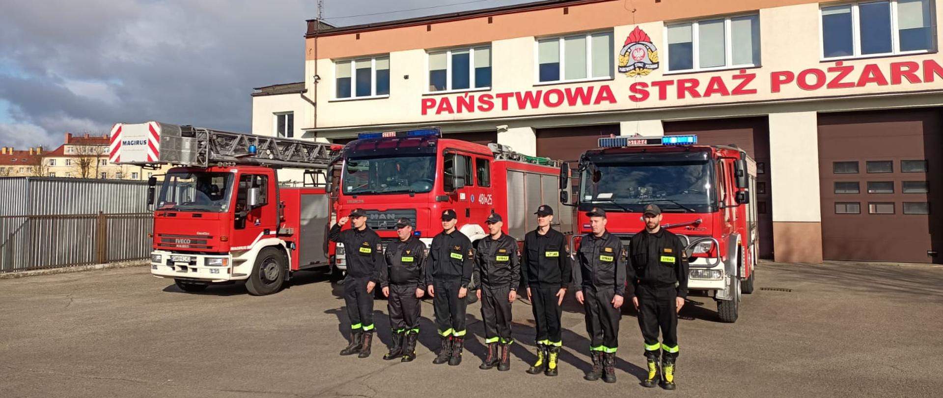 Fotografia przedstawia strażaków stojących na baczność w rzędzie, za nimi stoją samochody pożarnicze z włączonymi sygnałami świetlnymi koloru niebieskiego na tle budynku komendy powiatowej PSP w Malborku.