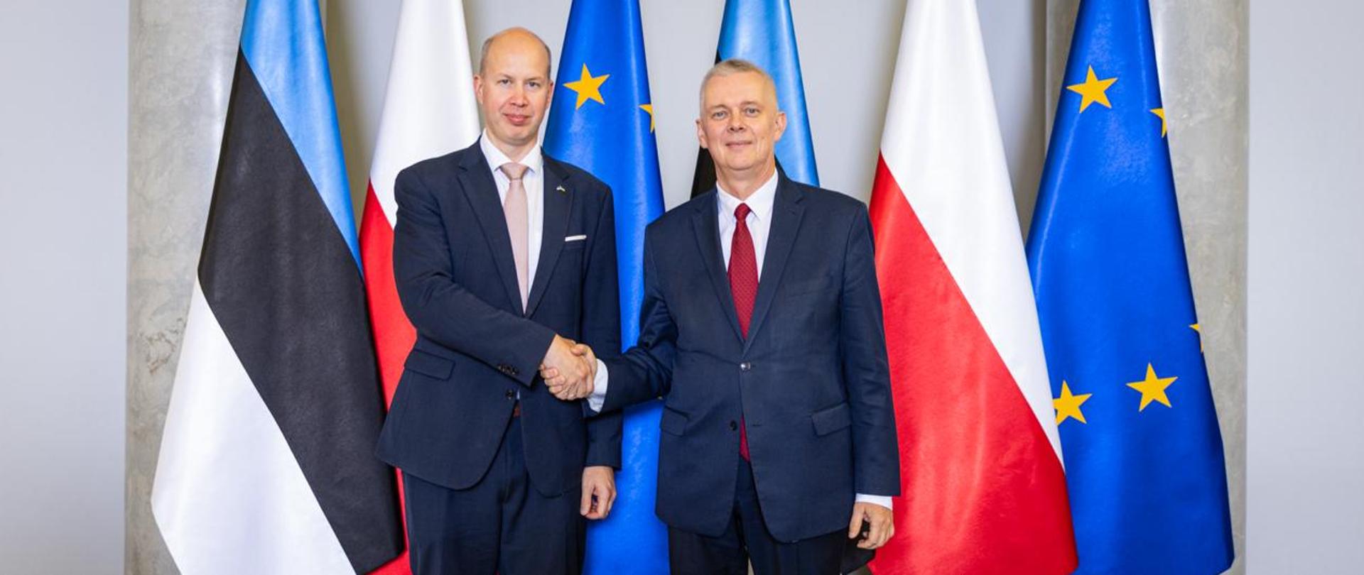 Minister Tomasz Siemoniak (z prawej) oraz minister Igor Taro stoją na tle sześciu flag. Flagi w konfiguracji od prawej: UE (niebieska z żółtymi gwiazdkami), Polski (biało-czerwona), Estonii (niebiesko-czarno-biała), UE, Polski, Estonii. W tle po prawej i lewej stronie widać dwie szare kolumny.