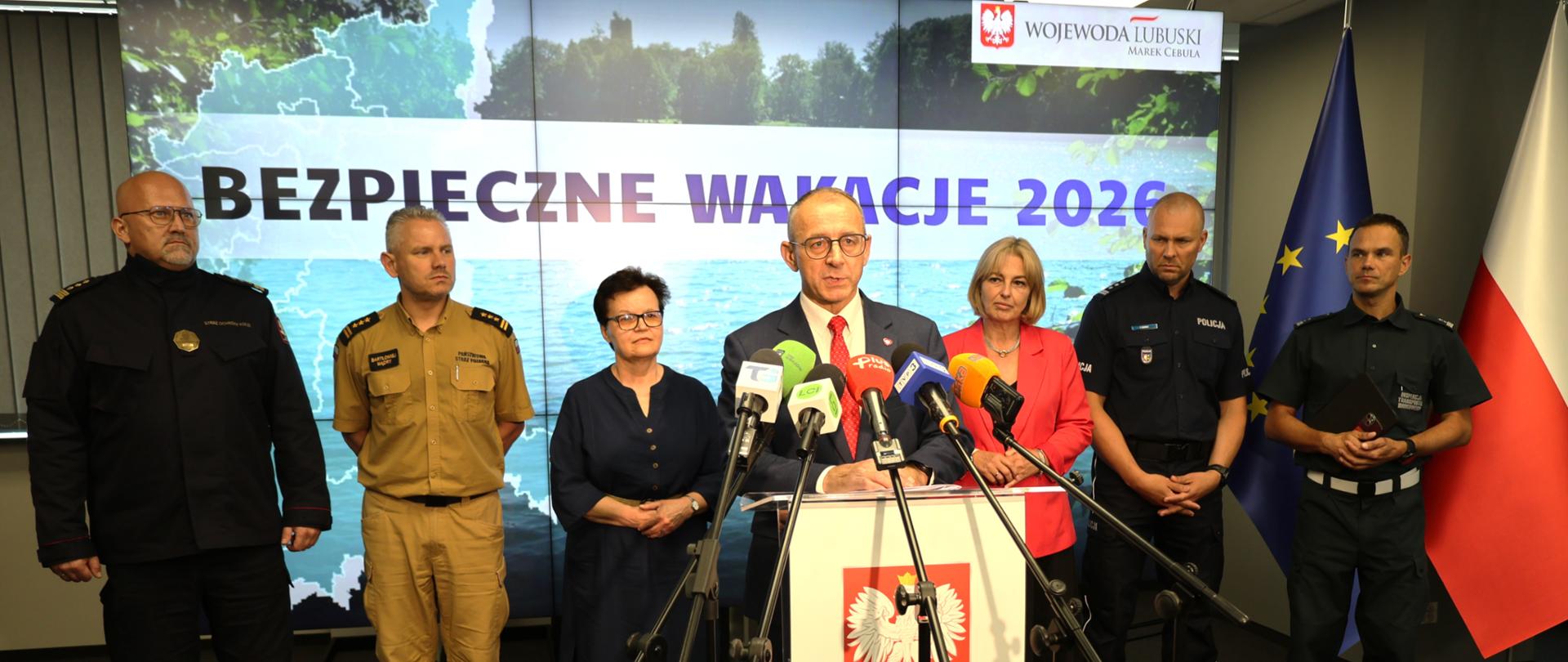 Bezpieczne wakacje 2026 - briefing prasowy. Marek Cebula Wojewoda Lubuski przemawia do mikrofonu. W tle przedstawiciele służb i instytucji oraz grafika "Bezpieczne wakacje".