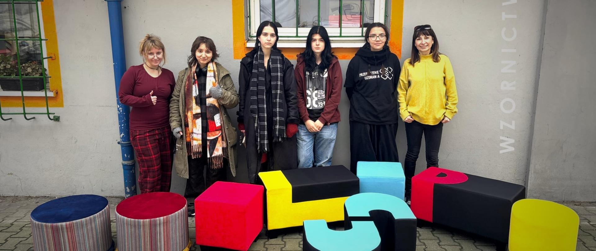 Na tle ściany z oknami stoi grupa sześciu osób, przednimi stoją zrealizowany w pracowni wzornictwa artystycznego pufy tapicerowane tworząca napis LSP