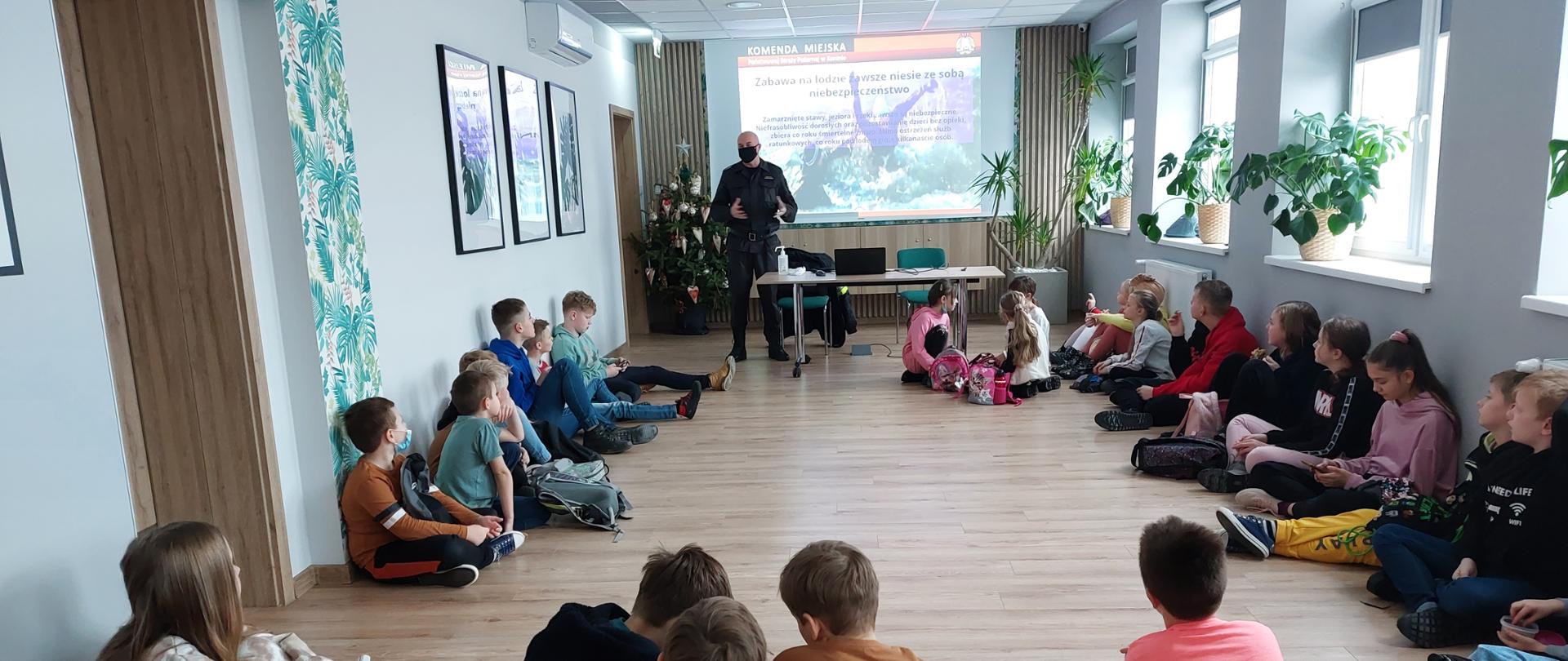 Zdjęcie przedstawia strażaka, który przeprowadza pogadankę dla dzieci z półkolonii zimowych. Dzieci siedzą wokół sali, a strażak stoi i informuje dzieci o zagrożeniach podczas ferii zimowych