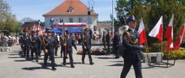 Zdjęcie przedstawia uroczystości o charakterze państwowym lub patriotycznym, odbywające się na placu w polskim mieście. Prawdopodobnie jest to święto narodowe (np. 3 Maja), na co wskazuje słoneczna pogoda i liczne polskie barwy narodowe.
Oto szczegóły widoczne na fotografii:
Uczestnicy i mundury
• Poczet sztandarowy i delegacje: Na pierwszym planie oraz w głębi widać grupy mężczyzn w ciemnogranatowych mundurach wyjściowych ze złotymi sznurami (akselbantami). Są to najprawdopodobniej przedstawiciele Straży Pożarnej (Ochotniczej lub Państwowej).
• Warta honorowa: Po prawej stronie maszeruje funkcjonariusz w pełnym umundurowaniu galowym, w rogatywce i czarnych rękawiczkach. W tle, przy pomniku, stoją żołnierze w mundurach polowych oraz beretach (prawdopodobnie Wojsko Polskie).
• Poczty sztandarowe: Widoczne są liczne sztandary niesione przez delegacje, co podkreśla oficjalny charakter wydarzenia.
Otoczenie i architektura
• Ratusz: W tle znajduje się charakterystyczny, biały budynek z wieżą zegarową, który pełni funkcję ratusza. Budynek jest udekorowany dużą biało-czerwoną flagą.
• Miejsce pamięci: W centralnej części placu widać pomnik, pod którym złożono liczne wieńce i wiązanki kwiatów w barwach narodowych.
• Symbole narodowe: Po prawej stronie ustawiony jest stojak z kilkoma polskimi flagami.
Atmosfera
• Wydarzenie odbywa się w pełnym słońcu na wybrukowanym placu miejskim.
• W tle, za barierkami, widać zgromadzonych mieszkańców obserwujących ceremonię. Obecne są również niebieskie namioty promocyjne (na jednym widnieje napis sugerujący nazwę miejscowości, prawdopodobnie Błonie).
Całość kompozycji oddaje podniosły i uroczysty nastrój lokalnych obchodów patriotycznych.
