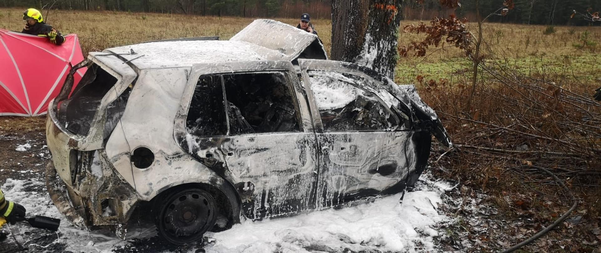 Zdjęcie przedstawia wrak spalonego samochodu, który wcześniej uderzył w drzewo. Auto pokryte jest pianą gaśniczą. Za autem znajduje się policjant, który rozmawia przez telefon oraz strażak trzymający czerwony parawan.