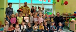 Obraz przedstawia zdjęcie zbiorowe, na którym znajdują się dzieci oraz dwóch strażaków. Zdjęcie ma miejsce w pomieszczeniu przedszkola, zielone ściany, piaskowo-zielone mebelki na których znajdują się zabawki oraz klocki. Za pozującymi widzimy okna z niebieskimi roletami. Strażak po prawej stronie kadru ubrany w czarne ubranie sztabowe na lewym ramieniu uwidocznione jest logo Państwowej Straży Pożarnej, widzimy także złote dystynkcje. Strażak stojący z lewej strony kadru ubrany jest w piaskowe ubranie specjalne z zielonymi poziomymi odblaskami, w prawej ręce trzyma czerwony hełm. 