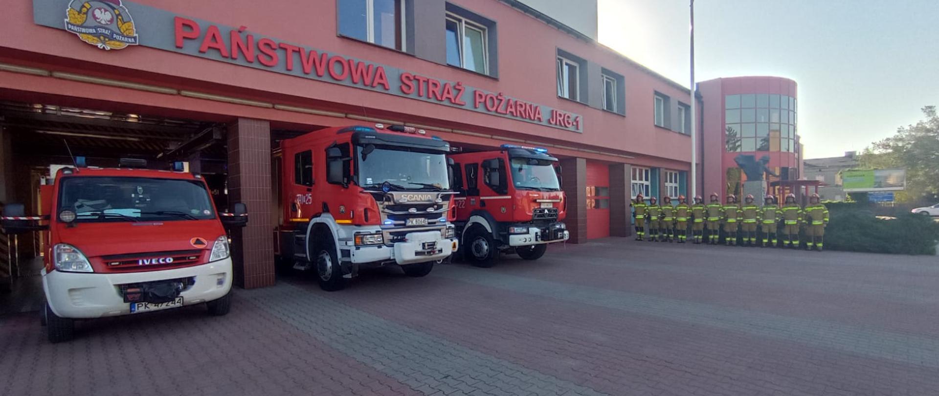 Na zdjęciu strażacy przed budynkiem strażnicy. Strażacy stoją przed garażami. Strażacy w umundurowaniu specjalnym w kolorze piaskowym. Zdjęcie wykonane w ciągu dnia. Strażacy posiadają czerwone hełmy. Za plecami stoją samochody ratowniczo-gaśnicze w kolorze czerwonym z sygnalizacją ostrzegawczą.