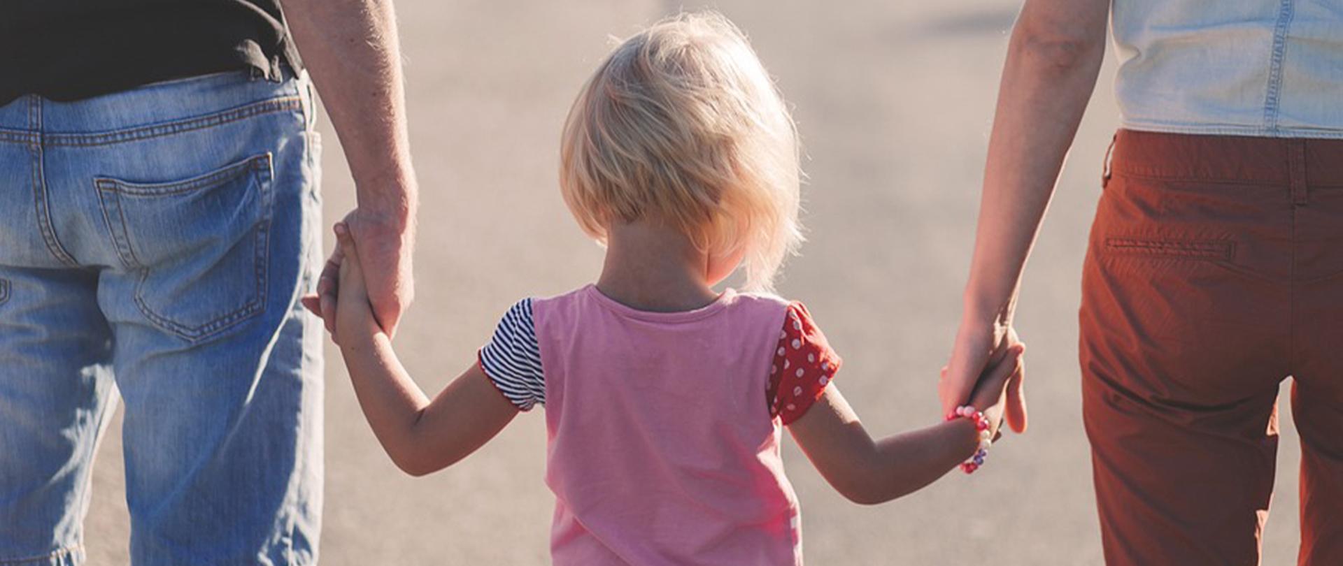 Na zdjęciu: dziecko trzymające za ręce dwie osoby