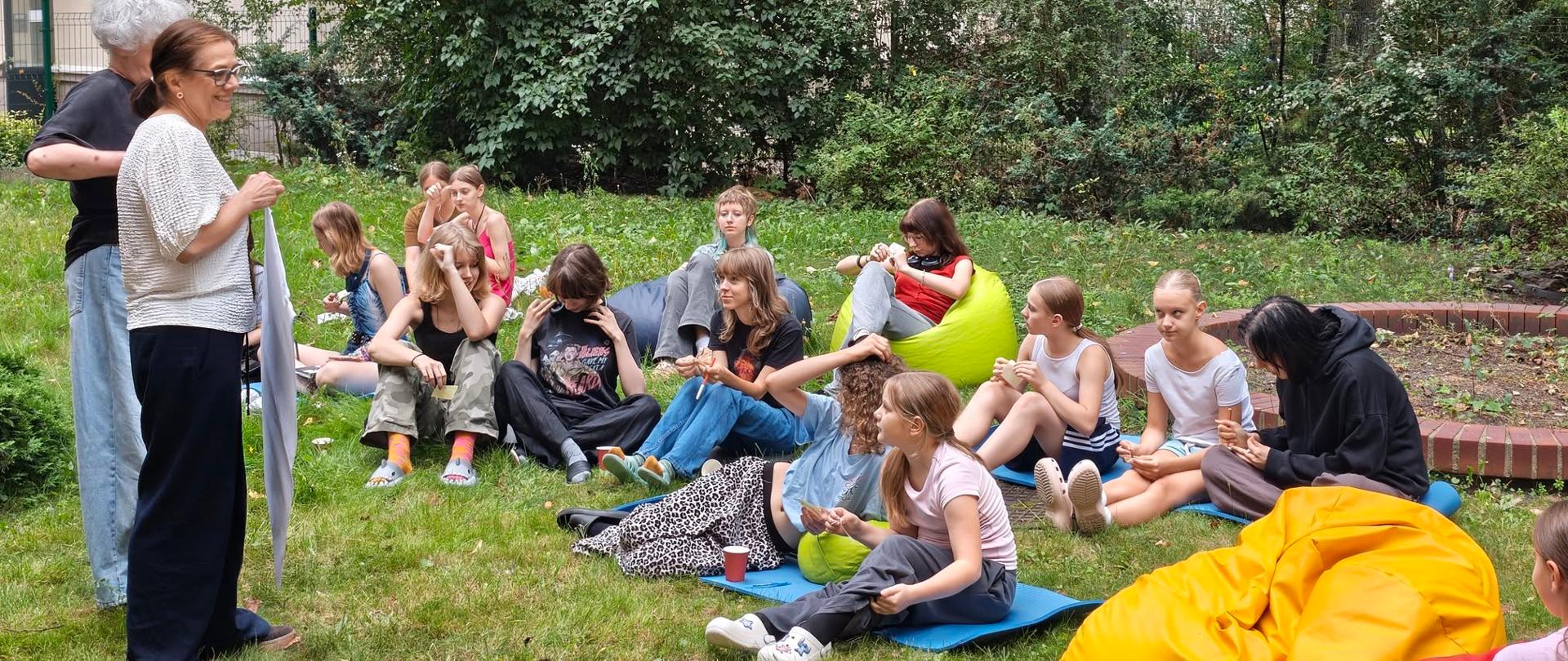 Grupa dziewcząt siedzi na kocach w bursianym ogródku, patrzą i słuchają instrukcji od pań wychowawczyń.