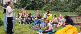Grupa dziewcząt siedzi na kocach w bursianym ogródku, patrzą i słuchają instrukcji od pań wychowawczyń.