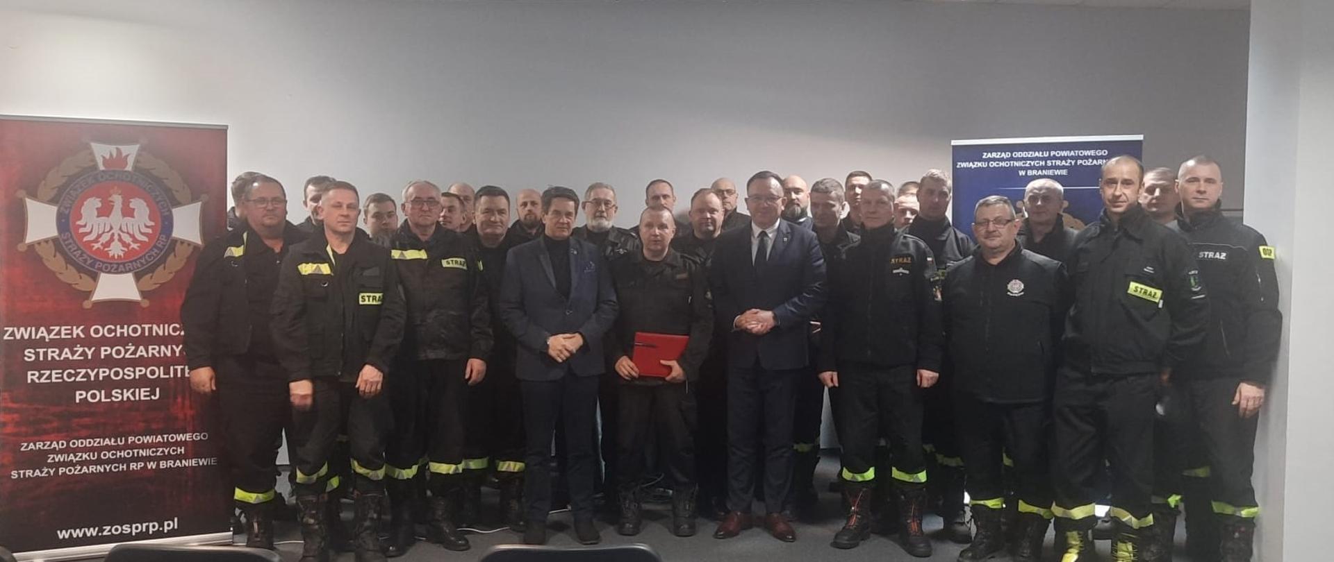 Na zdjęciu grupa kilkudziesięciu strażaków w czarnych mundurach oraz dwóch samorządowców w garniturach. Wszyscy stoją na tle białej ściany. Z lewej strony stoi baner z logiem i napisem Związek Ochotniczych Straży Pożarnych.