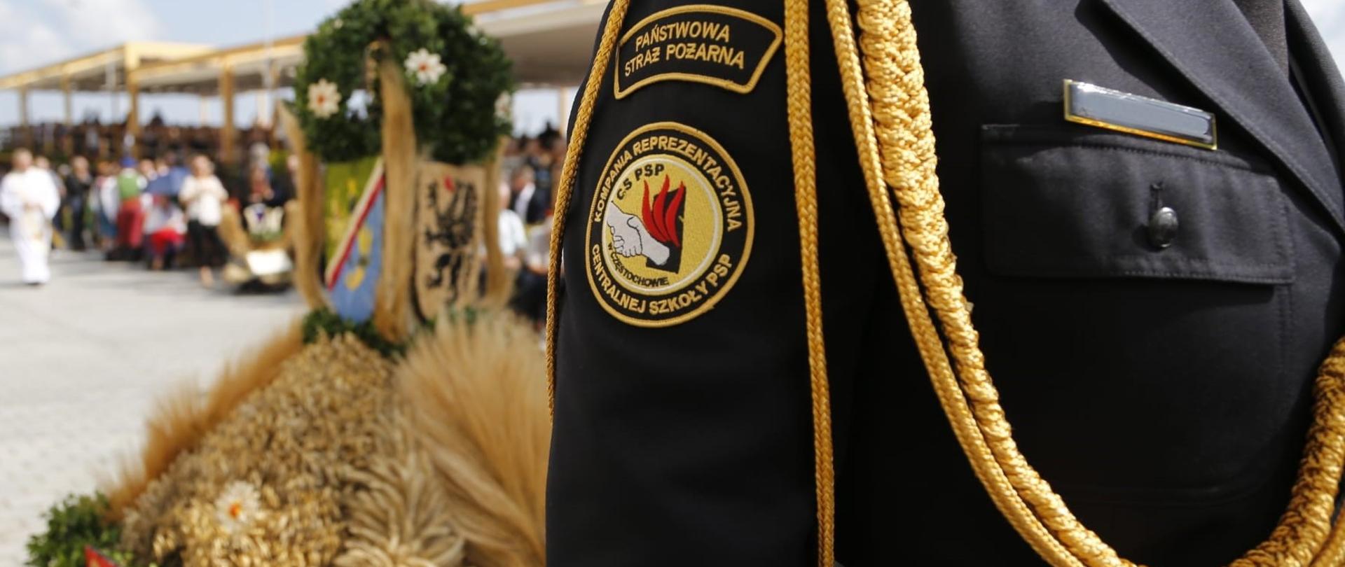 Na pierwszym planie ramię galowego munduru strażackiego z naszywką "Kompania reprezentacyjna Centralnej Szkoły PSP", w głębi kosz dożynkowy