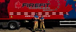 Na zdjęciu widzimy dwie osoby w spodniach strażackich i granatowych koszulkach stojące obok siebie z założonymi rękami. Za nimi znajduje się duża, czerwona naczepa z logo FIRE FIT Championships Europe oraz adresem strony internetowej „www.firefit-europe.de”
. Na grafice widać także sylwetkę strażaka oraz duży liść klonu z gwiazdami ułożonymi w okrąg.