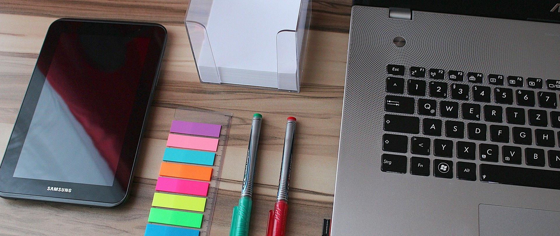 Na zdjęciu laptop, tablet, cienkopisy i karteczki samoprzylepne