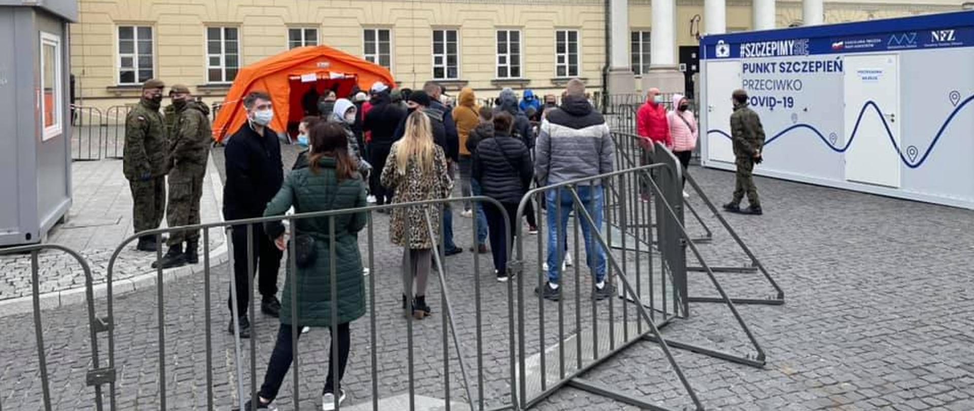 Na zdjęciu punkt szczepień oraz kolejka ludzi stojąca przy bramkach. 