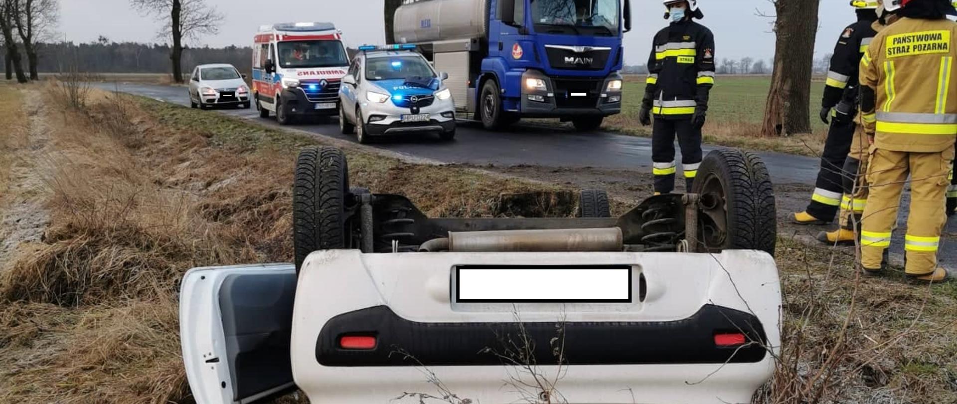 Zdjęcie przedstawia samochód osobowy koloru białego, który znajduje się na dachu w rowie. W tle radiowóz policji karetka pogotowia i strażacy.