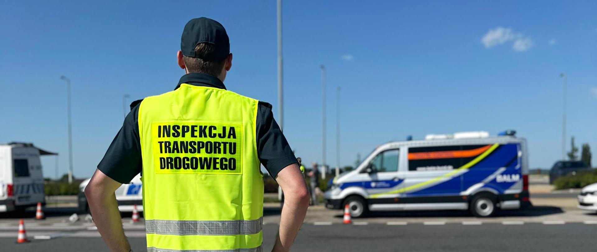 Funkcjonariusz w żółtej kamizelce z napisem Inspekcja Transportu Drogowego stoi tyłem. W tle widoczne są pojazdy służbowe i ciężarówki pod błękitnym niebem.