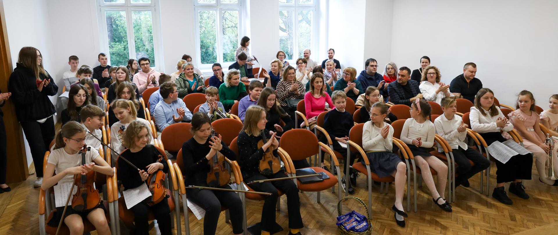 Siedemnasty Szkolny Konkurs Zespołów Kameralnych. Publiczność i wykonawcy zgromadzeni na auli PSM w Głubczycach. Widok ze sceny na publiczność, za oknami widać zazielenione drzewa.