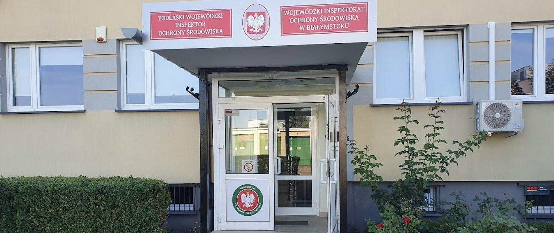 Ukazany budynek do urzędu na którym widać otwarte drzwi, schody, kostkę brukową i z obu stron zieleń.