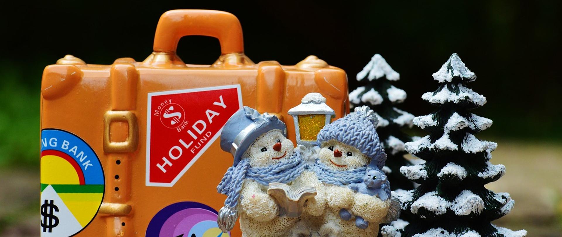 Przerwa świąteczne-zdjęcie przedstawia figurki bałwanków śnieżnych, ośnieżone drzewa i pomarańczową walizkę