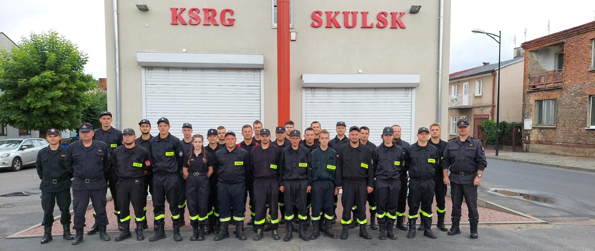 Zdjęcie przedstawia druhów OSP biorących udział w szkoleniu podstawowym, stojących na tle remizy z dwoma bramami garażowymi oraz napisem KSRG Skulsk.