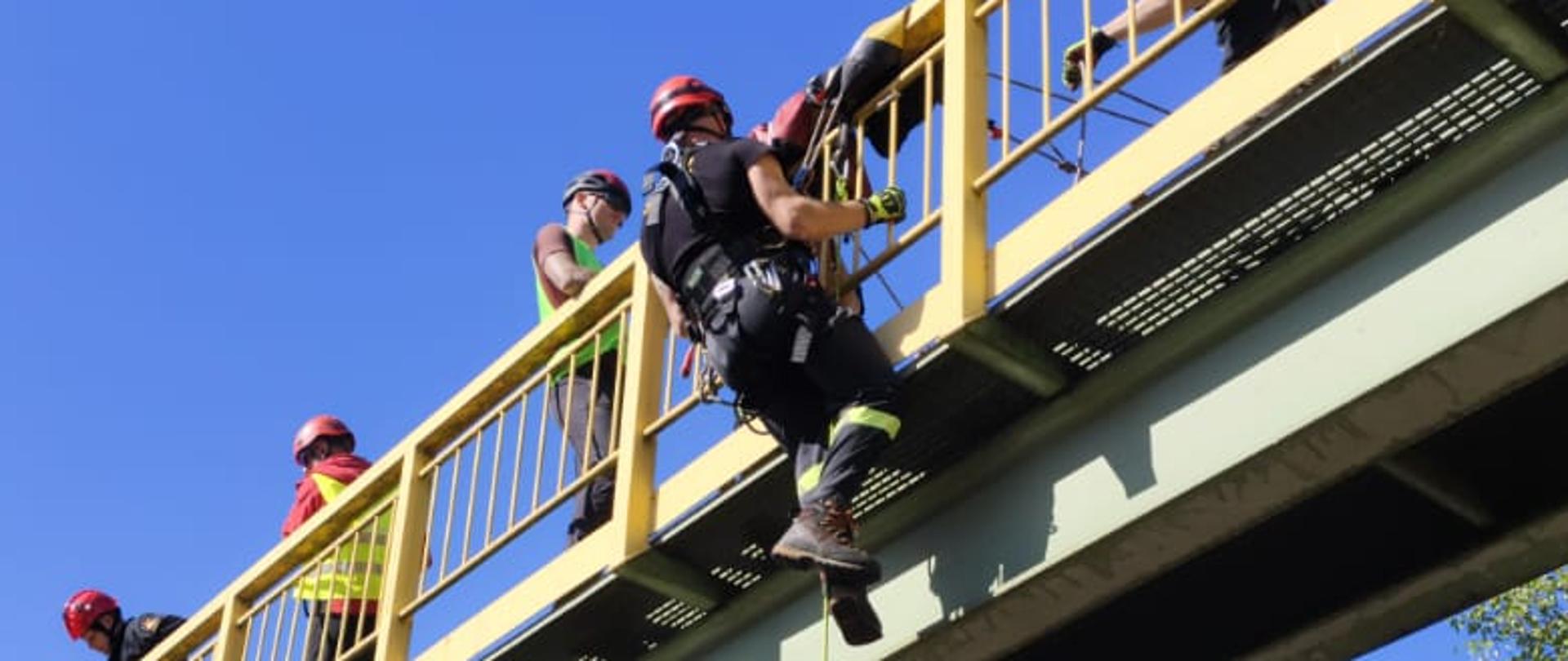 Działania na kładce dla pieszych. Osoba poszkodowana wisząca na lince nad nią ratownik wysokościowy schodzący do poszkodowanego. 