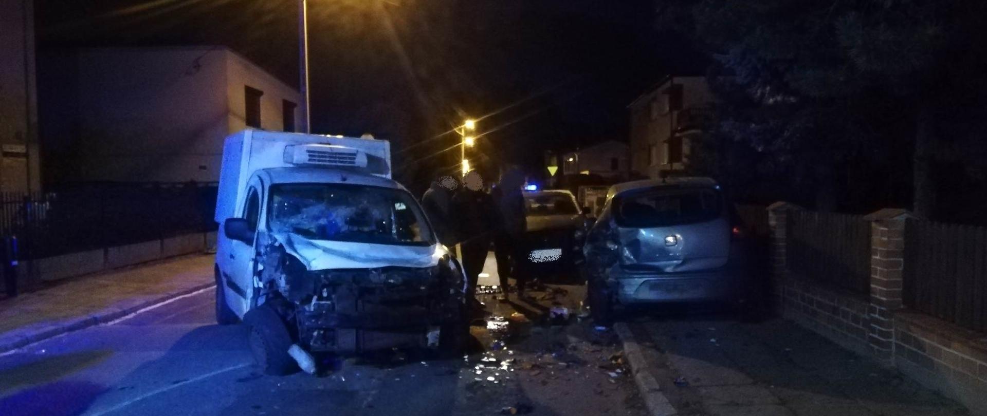 Zdjęcie przedstawia rozbite samochody biorące udział w zdarzeniu. Samochód dostawczy koloru białego ma uszkodzony przód, szary samochód osobowy ma uszkodzony tył. Przy pojazdach stoją właściciele środków transportu.