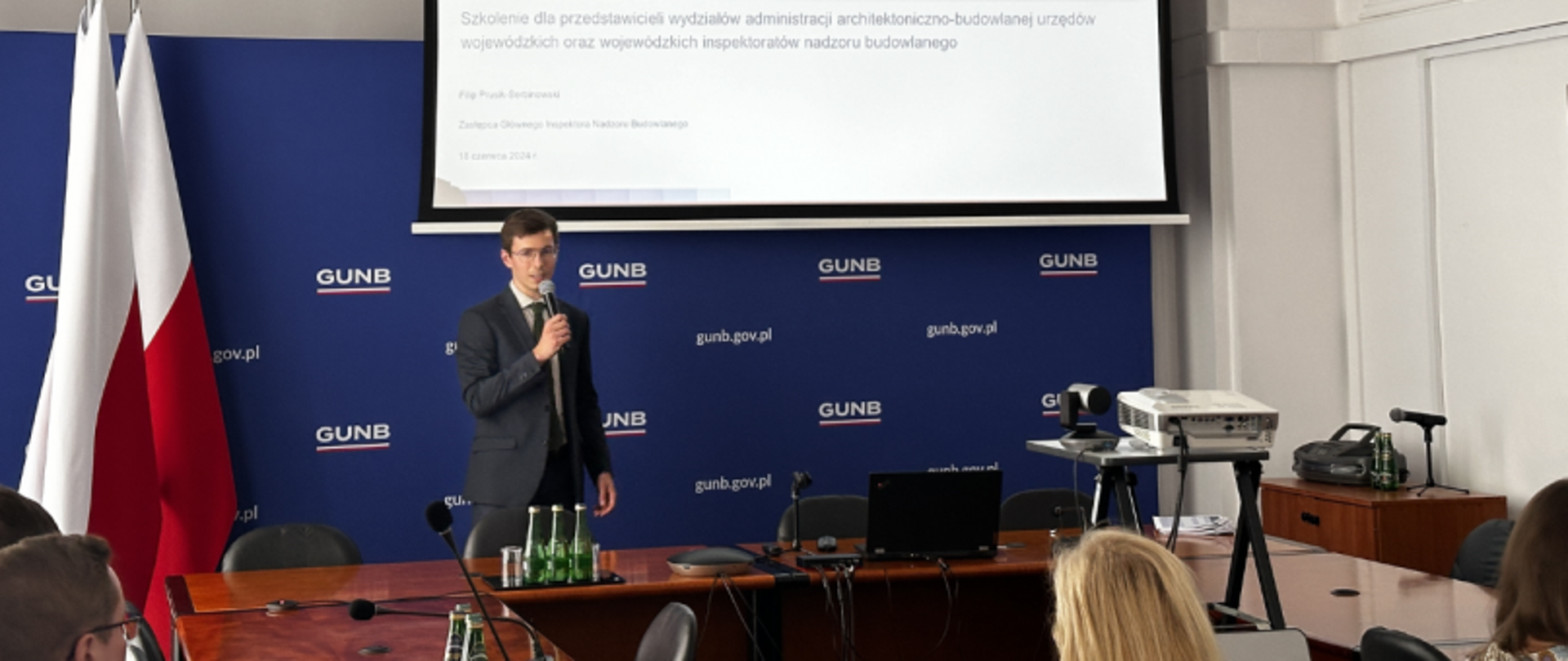 Szkolenie GUNB prowadzone przez Zastępcę Głównego Inspektora Nadzoru Budowlanego, z ekranem informacyjnym i logotypami w tle.