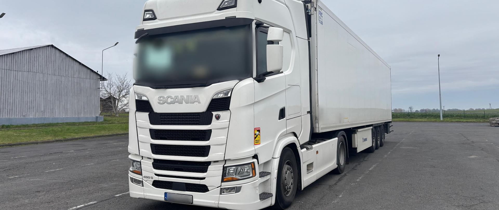 Biała ciężarówka marki Scania z naczepą chłodniczą stoi na asfaltowym parkingu na tle szarego, pochmurnego nieba. Kabina jest nowoczesna, masywna, z wyraźnymi liniami aerodynamicznymi i dużym grillem z logo „SCANIA” na przodzie. Naczepa jest długa, jednolicie biała, bez widocznych nadruków. W tle znajduje się niskie, szare budynki i fragment trawnika.