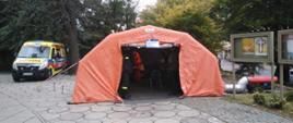 Na zdjęciu namiot koloru pomarańczowego po lewej stronie karetka pogotowia koloru żółtego w tle drzewa.