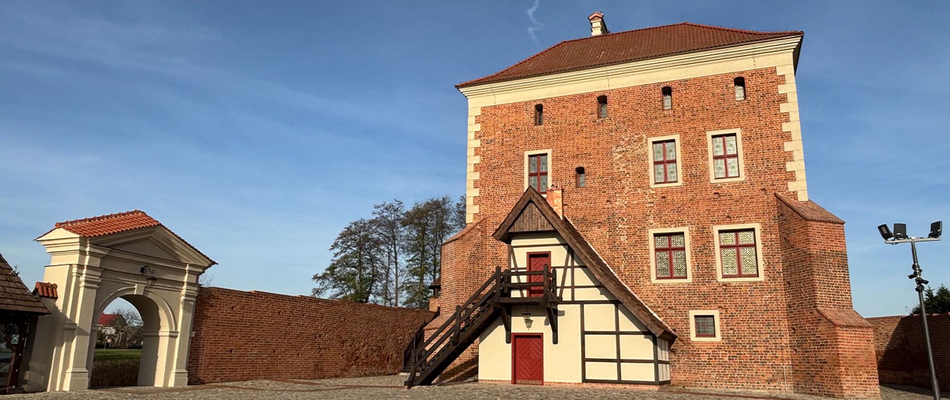 Zamek Kasztelański w Gołańczy po rewitalizacji. Na zdjęciu ceglana wieża donżonu, odtworzona przybudówka w konstrukcji szachulcowej ze schodami oraz brama i mur zamkowy na tle nieba.
