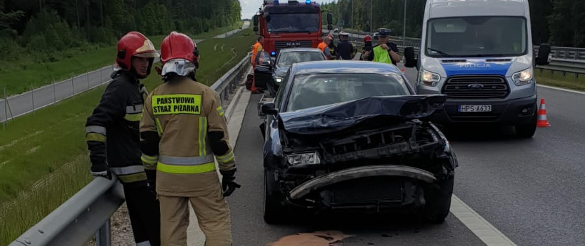 Na zdjęciu widać uszkodzony przód pojazdu marki Volkswagen po zderzeniu z barierą energochłonną. Przy pojeździe stoi dwóch strażaków. Za pojazdem widać radiowóz policyjny, oraz samochód pożarniczy, które mają włączone światła błyskowe.