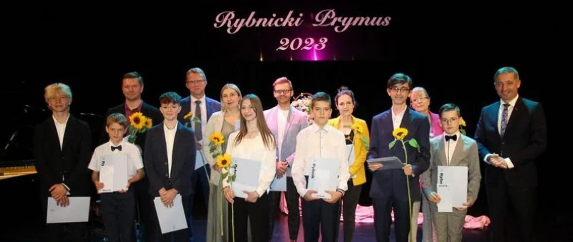 Kolorowe zdjęcie grupowe, na którym stoją w pierwszym rzędzie wyróżnieni uczniowie (& osób), w drugim nauczyciele (6 osób) i z prawej strony Prezydent Miasta Rybnika Piotr Kuczera. Wszyscy wyróżnieni trzymają w rękach dyplomy i słoneczniki.